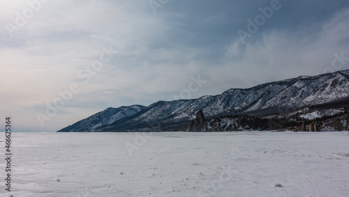 A wooded mountain range against a blue sky. A bizarre rock devoid of vegetation is visible on a frozen and snow-covered lake. Baikal