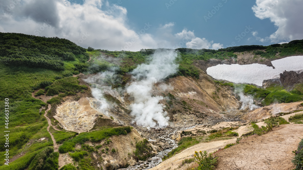 Smoke from fumaroles rises above the mountain slopes. Paths and areas of melted snow are visible. A stream from a hot spring flows along a rocky bed. Blue sky. Kamchatka