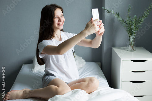 Portrait Of A Beautiful Brunette Teenager. The girl is sitting in her pajamas on her bed, looking out the window and smiling. Her Face Is Illuminated By The Warm morning sun. Beautiful room.