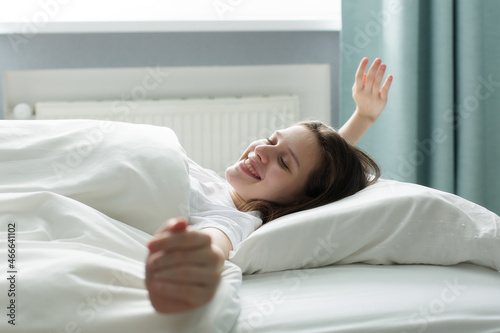 A beautiful teenager wakes up in the morning, stretches in bed, bright light on her from a large window. Happy teenage girl meets a new day in a beautiful room