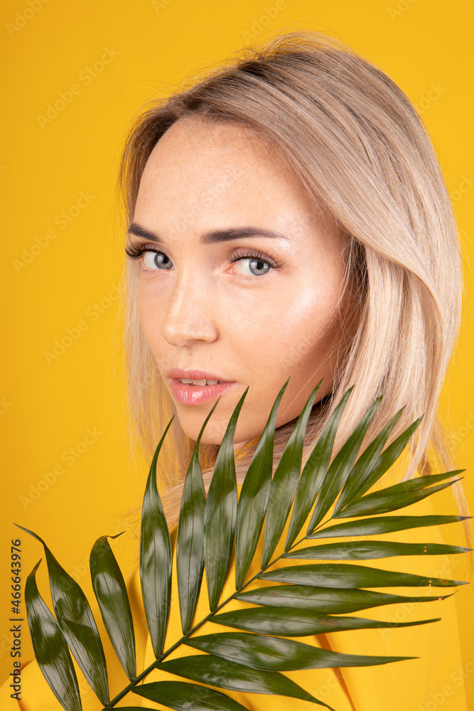 Naklejka premium Beauty portrait of a young, beautiful blonde woman with good skin in yellow clothes holding a green leaf on a yellow background