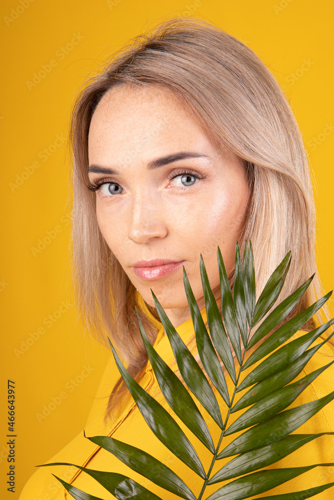 Naklejka premium Beauty portrait of a young, beautiful blonde woman with good skin in yellow clothes holding a green leaf on a yellow background