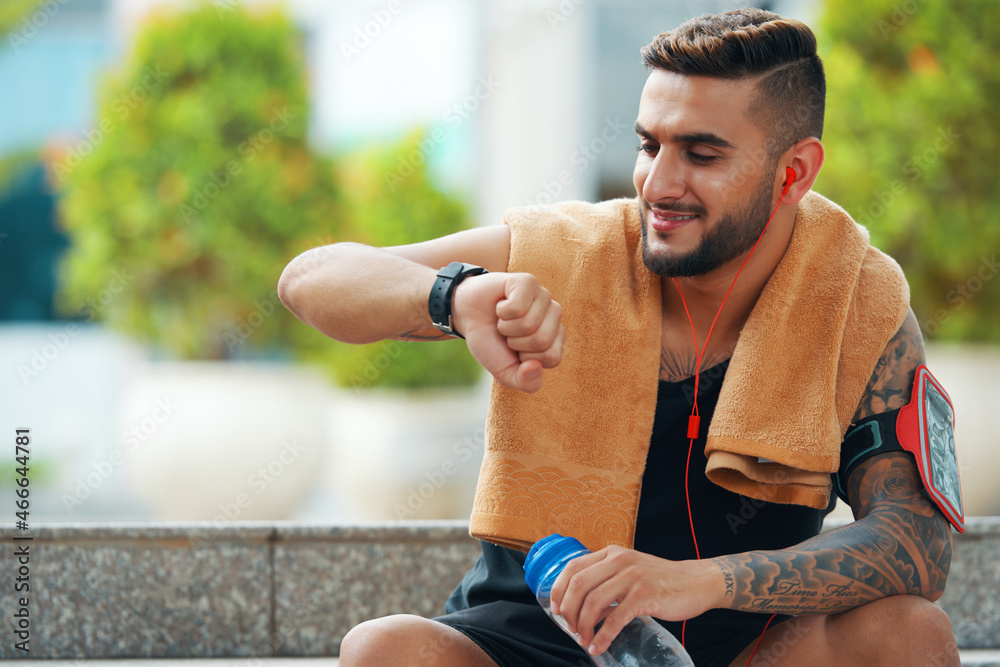 Smiling handsome young sportsman resting outdoors after training and checking fitness trekker Smiling handsome young sportsman resting outdoors after training and checking fitness trekker