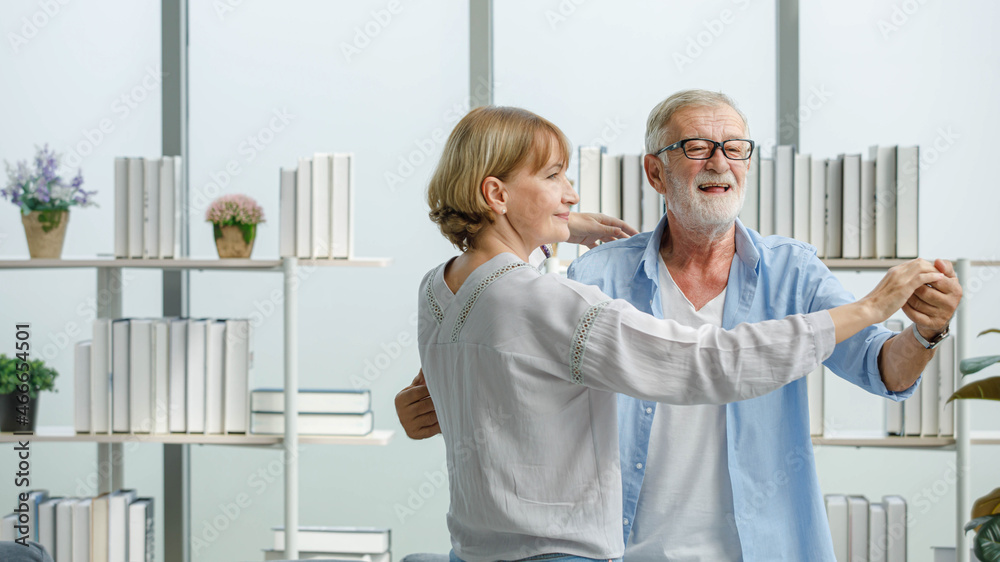 Obraz premium Caucasian old senior elderly happy grandparents lover married couple standing smiling look at each other face listening to slow waltz music dancing together in living room at home in front bookshelf