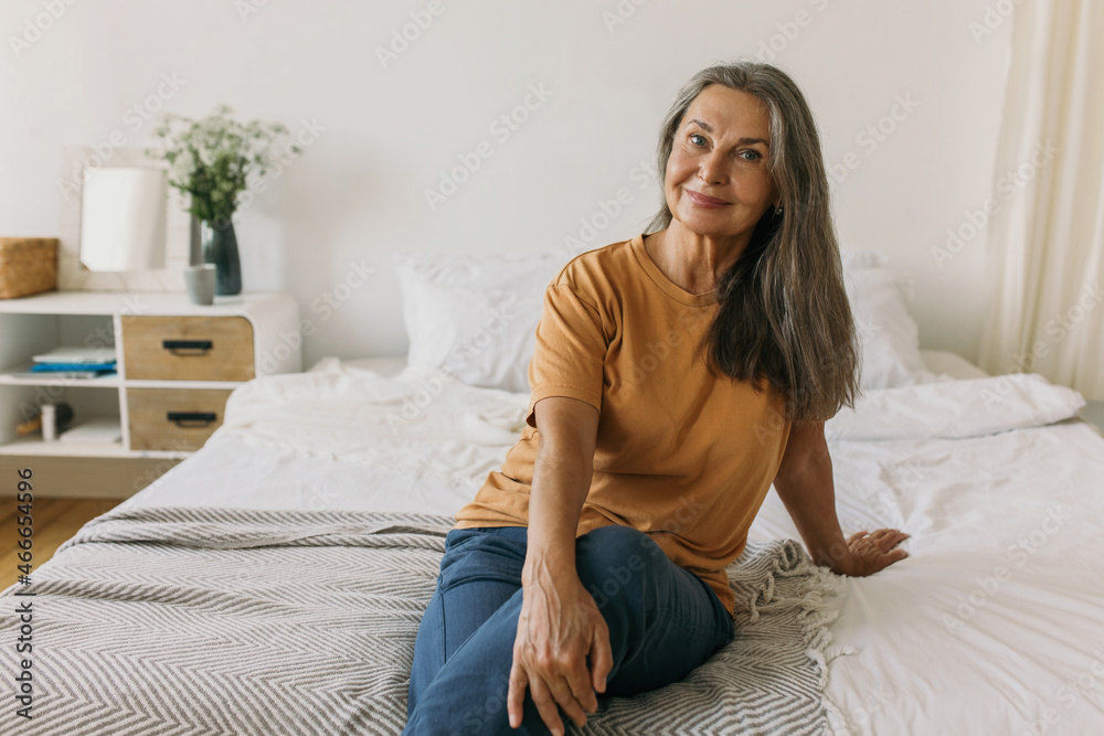 Pretty casual mature woman with long grey hair wearing jeans posing with  legs crossed in her bedroom, smiling at camera, against bed background,  feeling relaxed, having delightful face expression Photos | Adobe