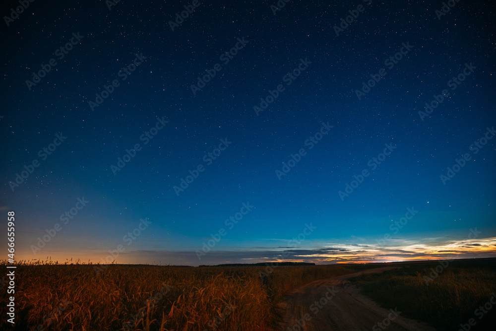 Night Starry Sky With Glowing Stars Above Countryside Landscape. Light Cloudiness Overcast Above Rural Field Meadow And Country Road In Summer