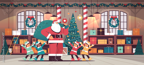 santa claus with mix race elves preparing for new year and christmas holidays celebration modern workshop interior