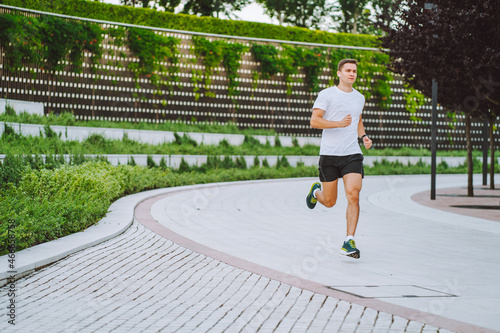 Attractive young man athlete wearing sportswear run in modern park at summer morning. Morning jogging, cardio, weight loss