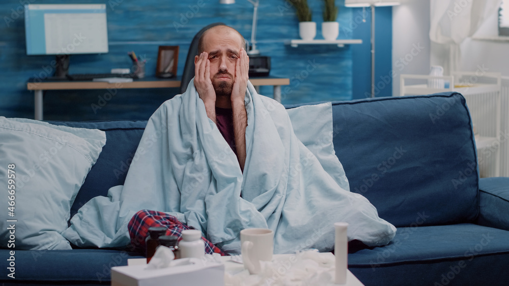 Sick man with headache and flu symptoms looking at camera while wrapped in blanket. Unhappy person feeling ill and rubbing temples against migraine. Adult with medicaments on table