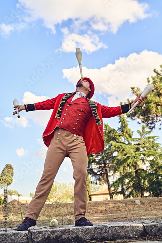 From below man in red costume balancing club on nose while juggling against cloudy blue sky