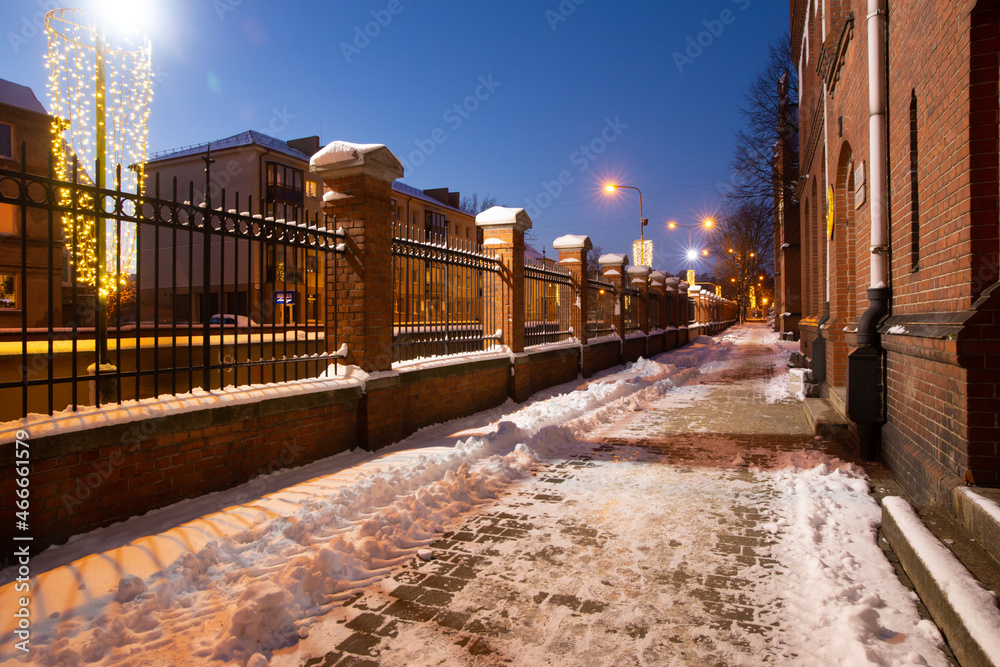 Klaipeda, Lithuania - 01 15 21: road near old historical red brick Neo-Gothic University building campus, the fence, winter snow, Christmas magic and city lights. An atmospheric photo for postcards