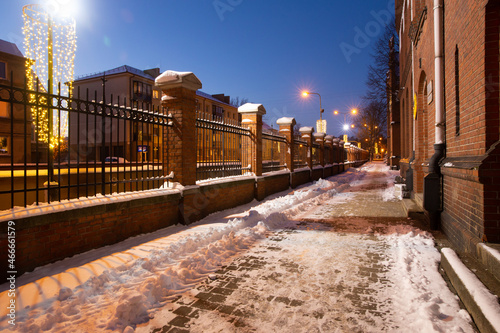Klaipeda, Lithuania - 01 15 21: road near old historical red brick Neo-Gothic University building campus, the fence, winter snow, Christmas magic and city lights. An atmospheric photo for postcards