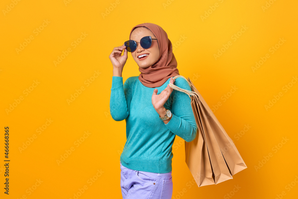 Cheerful young Asian woman wearing sunglasses and holding shopping bags on yellow background
