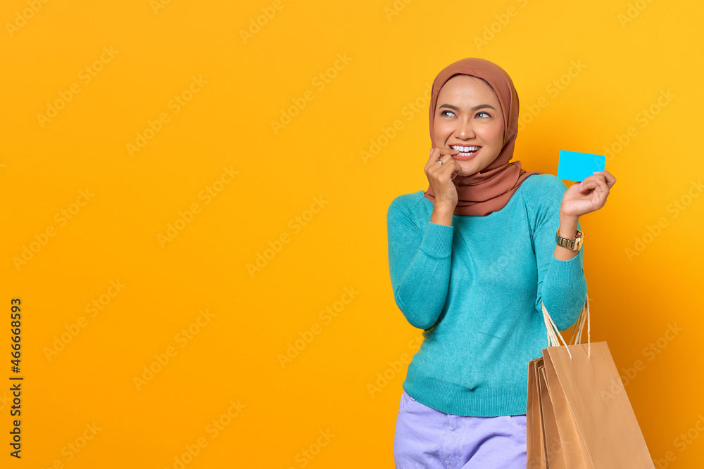 Smiling young Asian woman shopping holding credit card with a finger on lips on yellow background Smiling young Asian woman shopping holding credit card with a finger on lips on yellow background