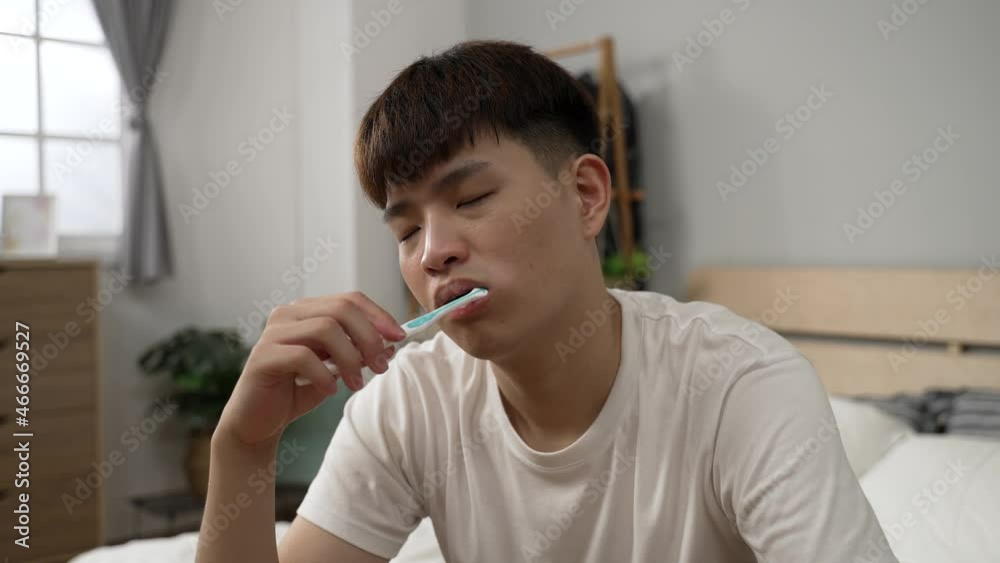 tired asian guy wearing t shirt is waking up from dozing off while brushing teeth in the morning in the bedroom at home. tired asian guy wearing t shirt is waking up from dozing off while brushing teeth in the morning in the bedroom at home.