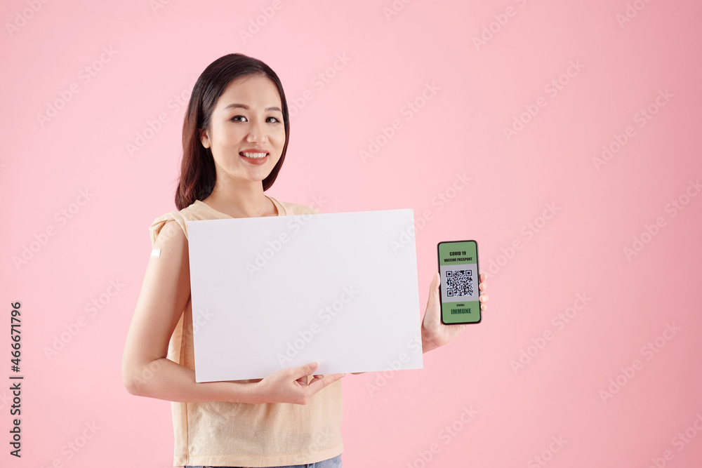 Smiling young woman holding white placard and smartphone with QR code proving that she was vaccinated Smiling young woman holding white placard and smartphone with QR code proving that she was vaccinated