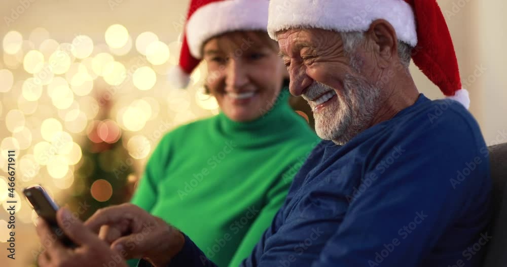 Smiling senior man with beard and mature daughter in Santa hats sitting on couch at home and chatting while browsing mobile phone together Smiling senior man with beard and mature daughter in Santa hats sitting on couch at home and chatting while browsing mobile phone together