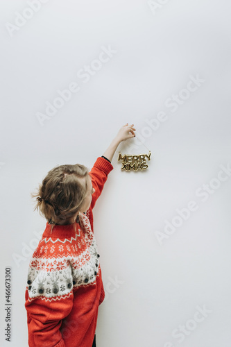 a modern woman in a red sweater hangs a Christmas inscription on a white wall. MERRY XMAS. The minimum concept of the new year