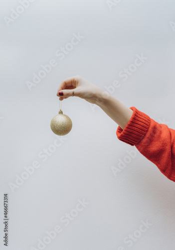 a woman 's hand in a red sweater holds a golden Christmas tree toy in the form of a ball , on a light background