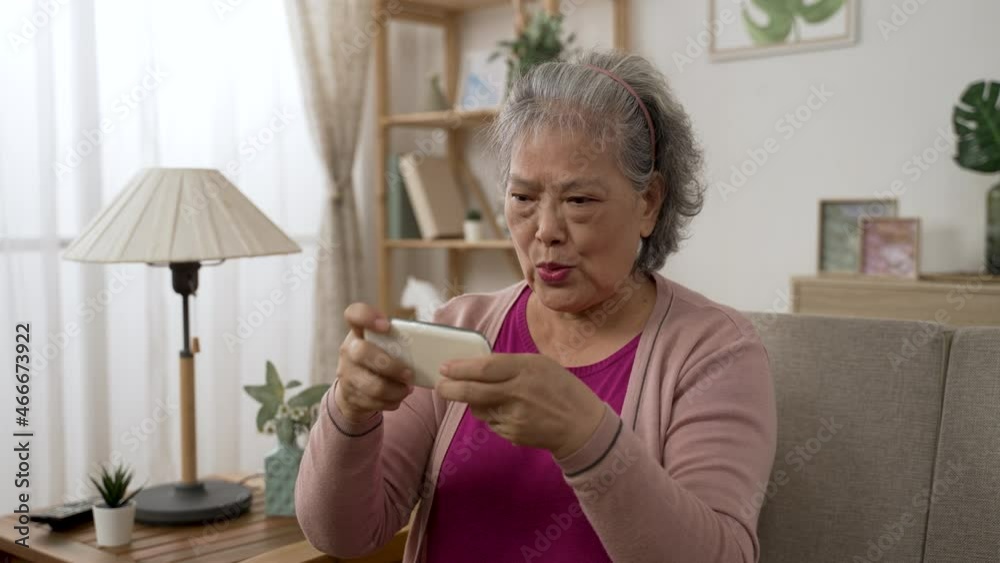 overjoyed asian elderly female player is lifting her hands to celebrate for winning exciting mobile game on the phone on the couch in the living room at home overjoyed asian elderly female player is lifting her hands to celebrate for winning exciting mobile game on the phone on the couch in the living room at home