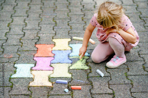 Little preschool girl painting with colorful chalks on ground on backyard. Positive happy toddler child drawing and creating pictures. Creative outdoors activity in summer.