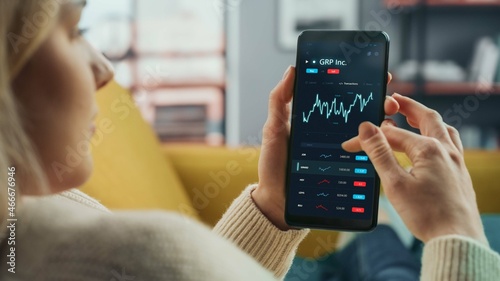 Young Woman at Home Lying on a Couch and Using Smartphone with Stock Market App to Check Share Prices. Graph is Going Up Making a Great Profit. In the Background Cozy Homely Atmosphere.