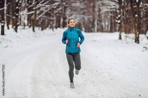Fit sportswoman running in nature on a snowy chilly winter day. Cold weather, snow, healthy life, fitness, forest