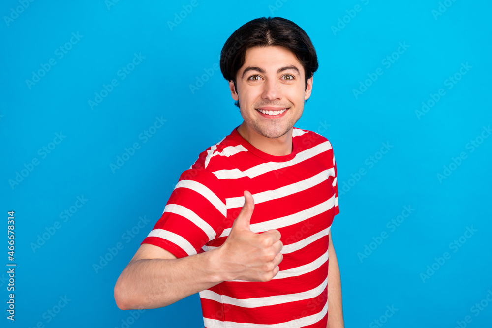 Photo of funky charming young man wear striped t-shirt smiling showing thumb up isolated blue color background Photo of funky charming young man wear striped t-shirt smiling showing thumb up isolated blue color background