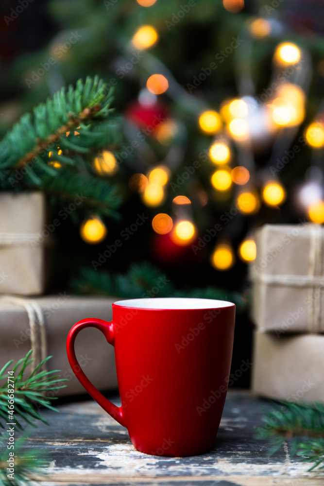 Coffee in a red cup on the rustic table with wrapped gifts, Christmas tree lights and spruce branches in the background Coffee in a red cup on the rustic table with wrapped gifts, Christmas tree lights and spruce branches in the background