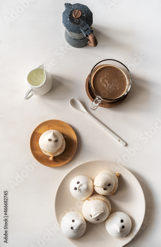 Meringues in a form of ghosts on the plate with cup of coffee, milk jar and coffe pot on the light background, flat lay.
