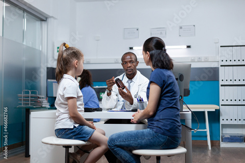 African american pediatrician doctor holding pills bottle explaining medicine prescription to mother during clinical appointment in hospital office. Therapist man discussing antibiotics treatment