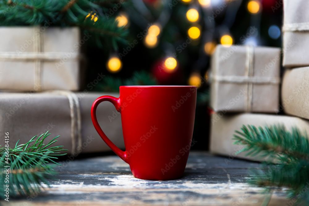 Coffee in a red cup on the rustic table with wrapped gifts, Christmas tree lights and spruce branches in the background Coffee in a red cup on the rustic table with wrapped gifts, Christmas tree lights and spruce branches in the background
