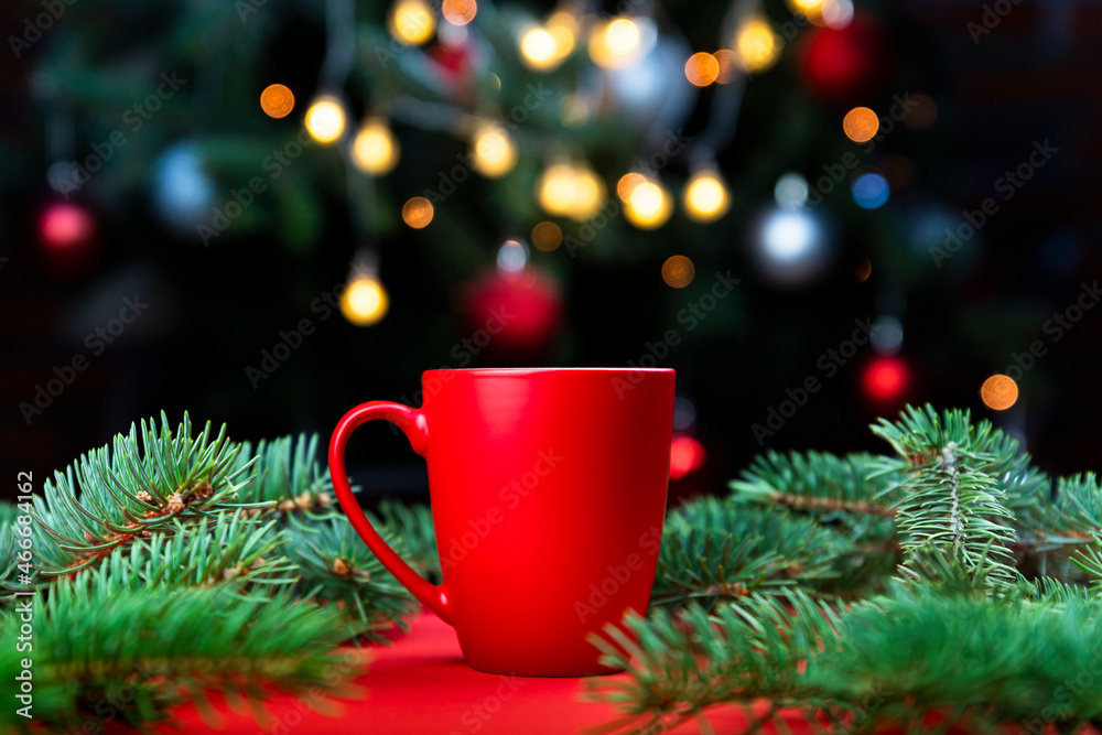 Morning coffee in a red cup on the red surface with Christmas tree lights and spruce branches in the background Morning coffee in a red cup on the red surface with Christmas tree lights and spruce branches in the background