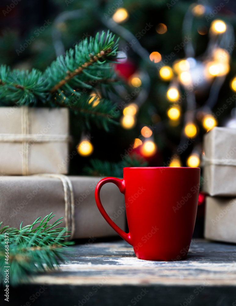 Coffee in a red cup on the rustic table with wrapped gifts, Christmas tree lights and spruce branches in the background