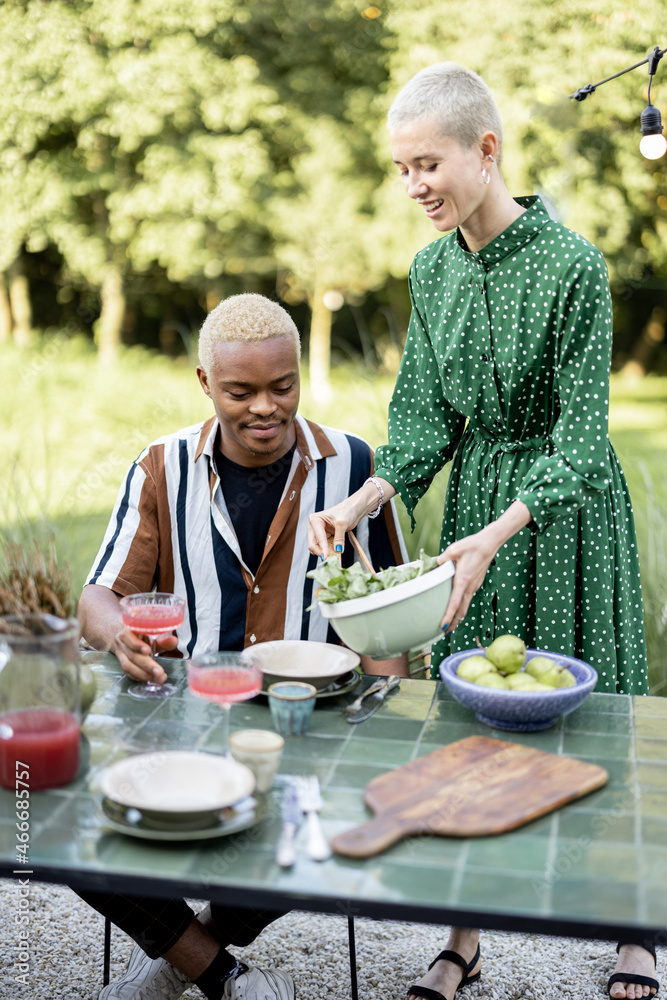 European woman putting salad on plate of her black boyfriend during dinner outdoors. Concept of relationship. Modern lifestyle. Couple enjoying time together. Idea of healthy eating. Sunny daytime