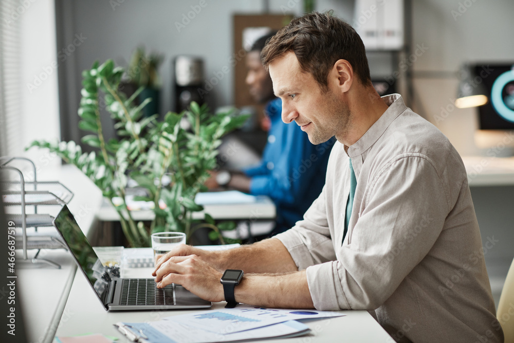 Side view portrait of smiling adult man using laptop while enjoying work in office, copy space - obrazy, fototapety, plakaty 