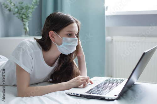 a teenage schoolgirl in a mask, a student of a virtual remote online digital class, looks at a laptop while sitting on a bed at home. stop coronavirus