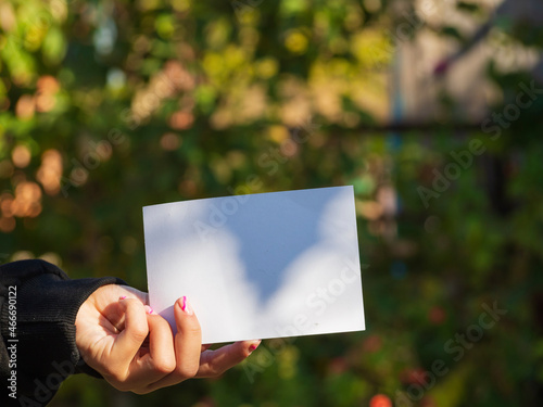 Copy space of white paper against the background of nature. Use it to place text on an empty space. A Girl Holds A Clean White Cardboard In Her Hands Against The Background Of A Blurred Green Garden