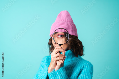 Cozy portrait of a young woman in a knitted blue sweater and a pink hat with bright makeup holding a magnifying glass, fooling around, having fun photo