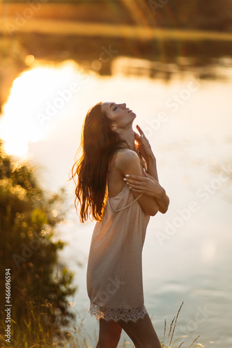 a young beautiful girl, of model appearance, in a shirt, stands among the landscape on the river bank, the setting sun illuminates her figure