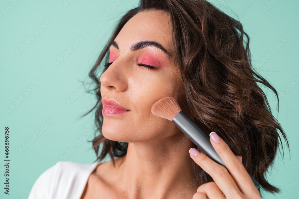 Young woman in a white evening dress on a light green background with bright pink makeup holding a blush brush Young woman in a white evening dress on a light green background with bright pink makeup holding a blush brush