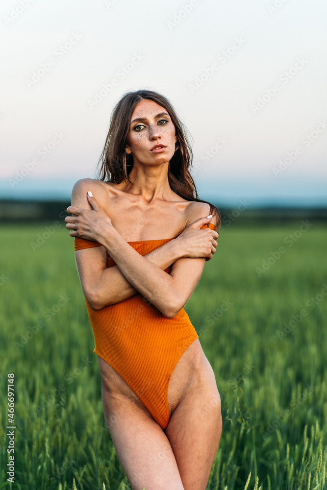 a young beautiful girl of model appearance in a bright orange bodysuit stands in the middle of a lush green field. a young beautiful girl of model appearance in a bright orange bodysuit stands in the middle of a lush green field.