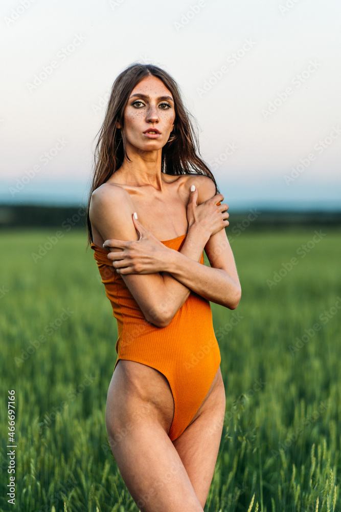 a young beautiful girl of model appearance in a bright orange bodysuit stands in the middle of a lush green field. a young beautiful girl of model appearance in a bright orange bodysuit stands in the middle of a lush green field.