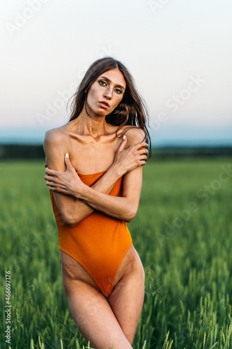 a young beautiful girl of model appearance in a bright orange bodysuit stands in the middle of a lush green field.