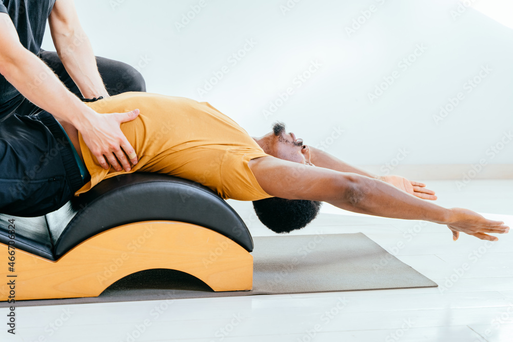 Close up of male instructor consulting handsome african european beard man workout pilates on small barrel practice in pilates studio, working out indoor, correcting beginners. Close up of male instructor consulting handsome african european beard man workout pilates on small barrel practice in pilates studio, working out indoor, correcting beginners.