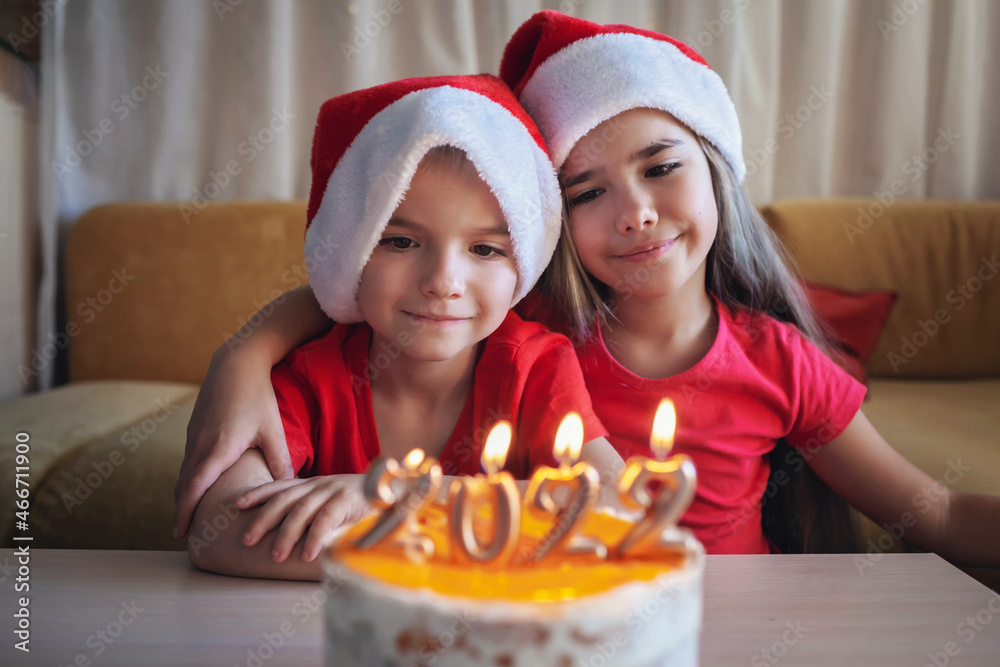Siblings in Santa hats look dreamily at burning candles on Christmas cake with golden 2022 numbers on top with yellow icing over bokeh lights background, family New Year celebration, focus on kids Siblings in Santa hats look dreamily at burning candles on Christmas cake with golden 2022 numbers on top with yellow icing over bokeh lights background, family New Year celebration, focus on kids