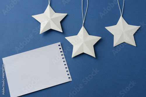 Three white paper stars and a blank sheet from a notebook on a dark blue background  top view. Children s  holiday or Christmas background with stars  copy space