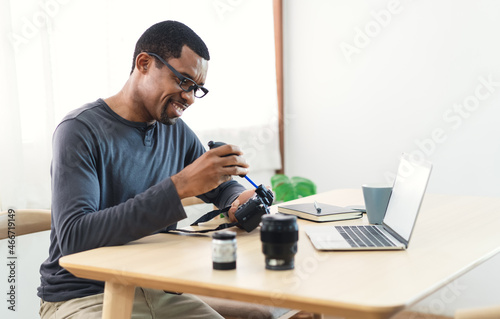 Handsome African photographer with her professional camera and cleaning the camera and lens while sit on desk at his studio. Freelance photographer online marketing. Creative artist lifestyle.