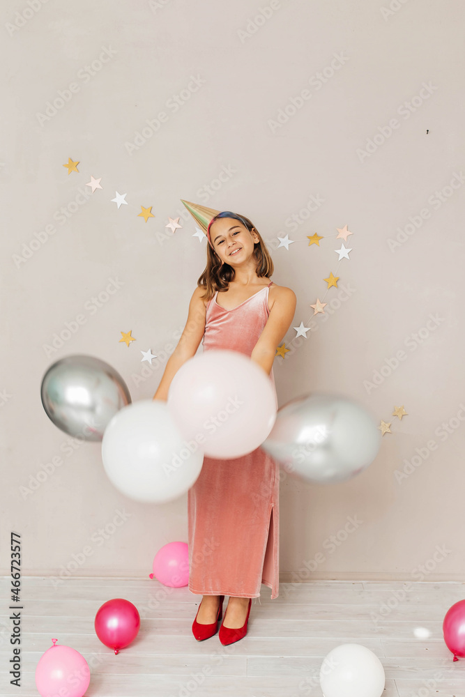 Fototapeta premium A beautiful teenager girl in an elegant pink dress, a festive cap and balloons celebrates her birthday.