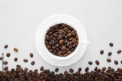 White cup with brown coffee beans on light gray table background. Closeup. Top down view.